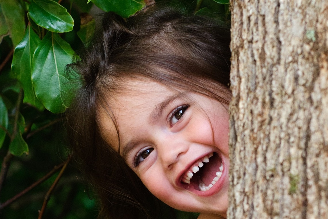 A young, smiling girl with brown hair pokes her head out from behind the trunk of a tree, with green leaves above her head.