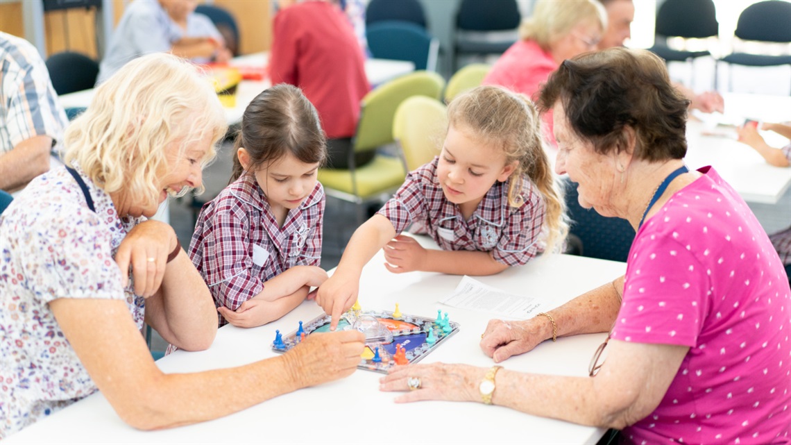 Two older women play a board game with 2 young schoolchildren in a well-lit classroom.