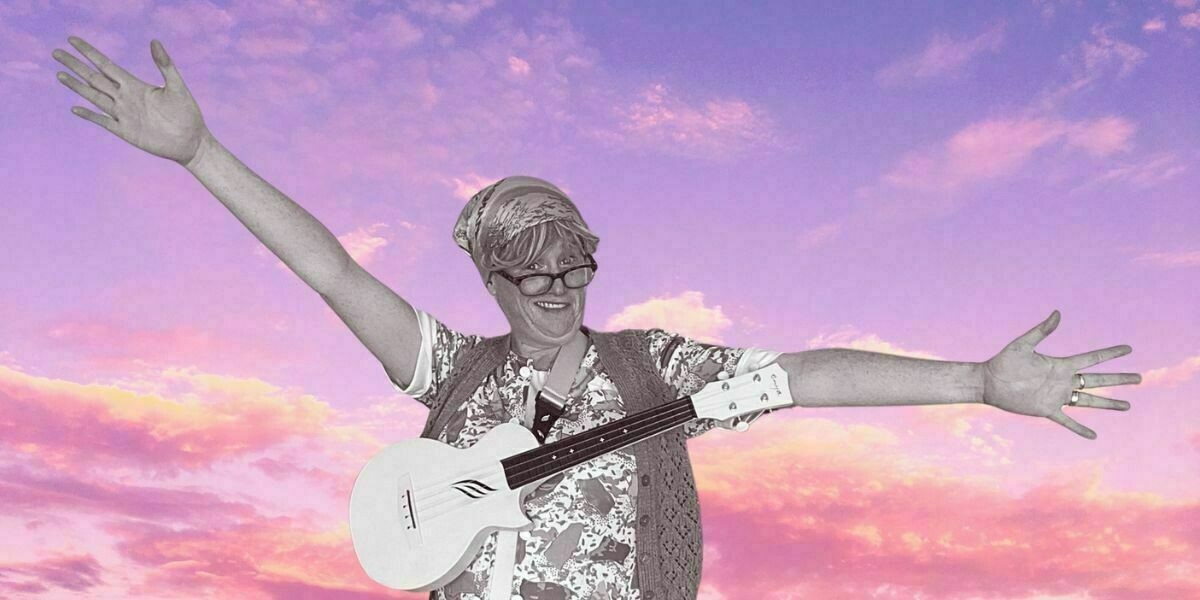 A smiling performer dressed as a granny smiles with her arms outstretched and a ukulele around her neck.