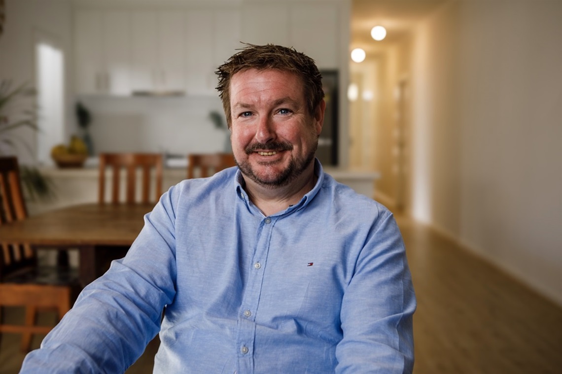 A man in a light blue shirt smiles towards the camera in the well-lit dining area of his Port Noarlunga South home.