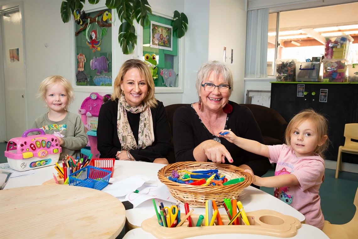 Two smiling young children sit either side of Junction's CEO and Onkaparinga Mayor Moira Were, with a table filled with toys, pencils and paper in front of them.
