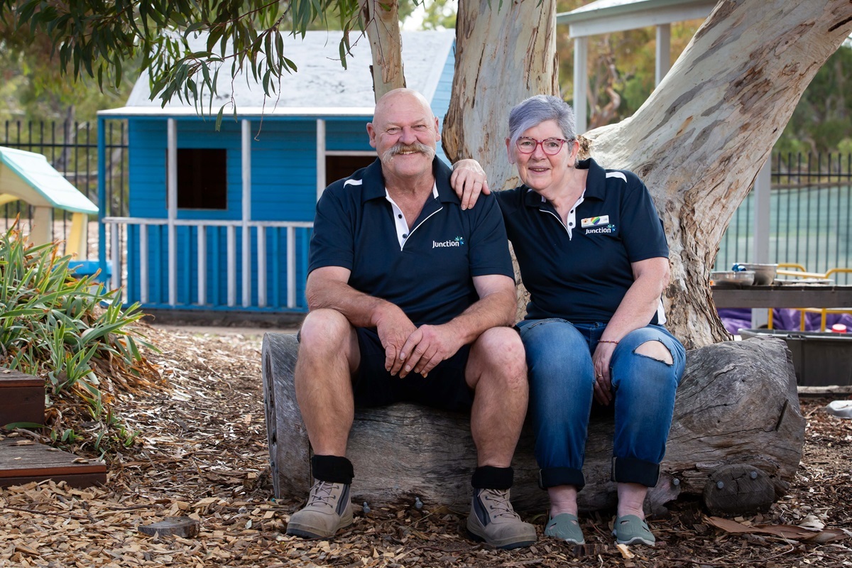 Husband and wife volunteer duo Len and Lou turner sit on a log at Hackham Community Centre and smile at the camera.