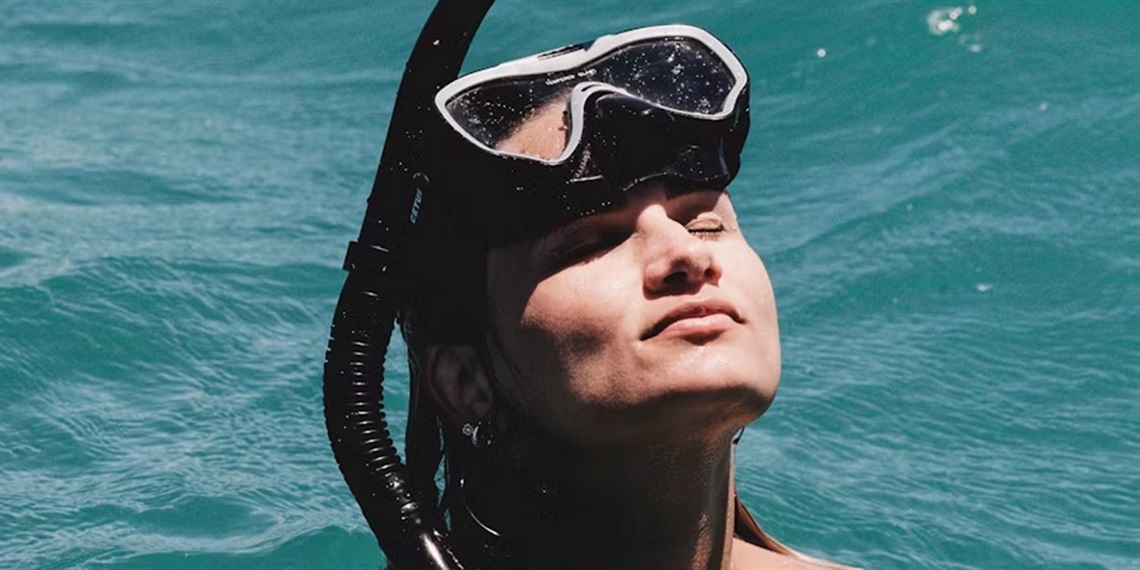 A close-up photo of a woman's face emerging from the ocean. Her eyes are closed, head pointing to the sun, and she has a black snorkel mask resting on her forehead.