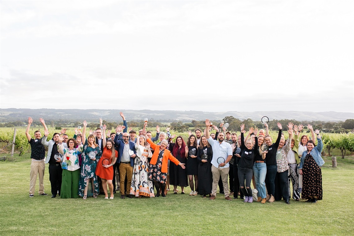 A large crowd shot of the 2025 Onkaparinga Business Award winners cheering with their awards on the green grass of a winery.