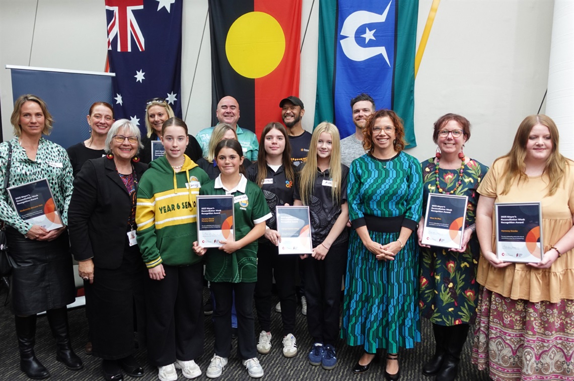 A group of smiling award recipients stand with the Onkaparinga Mayor and Amanda Rishworth MP holding their awards with the Aboriginal,Torres Strait Islander, and Australian flags behind them.