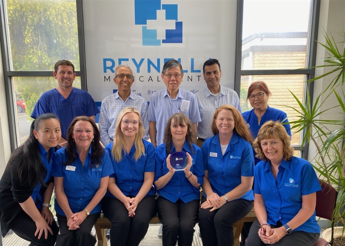 The team at Reynella Medical Centre smile in their blue uniforms with a woman in the middle holding their award.
