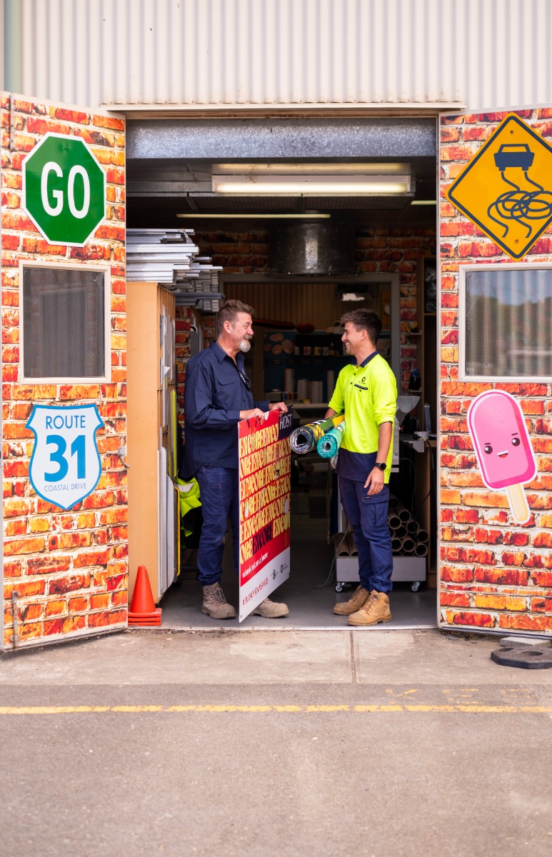 Two smiling Sign Shop workers talk between the colourful doorways of the Sign Shop.