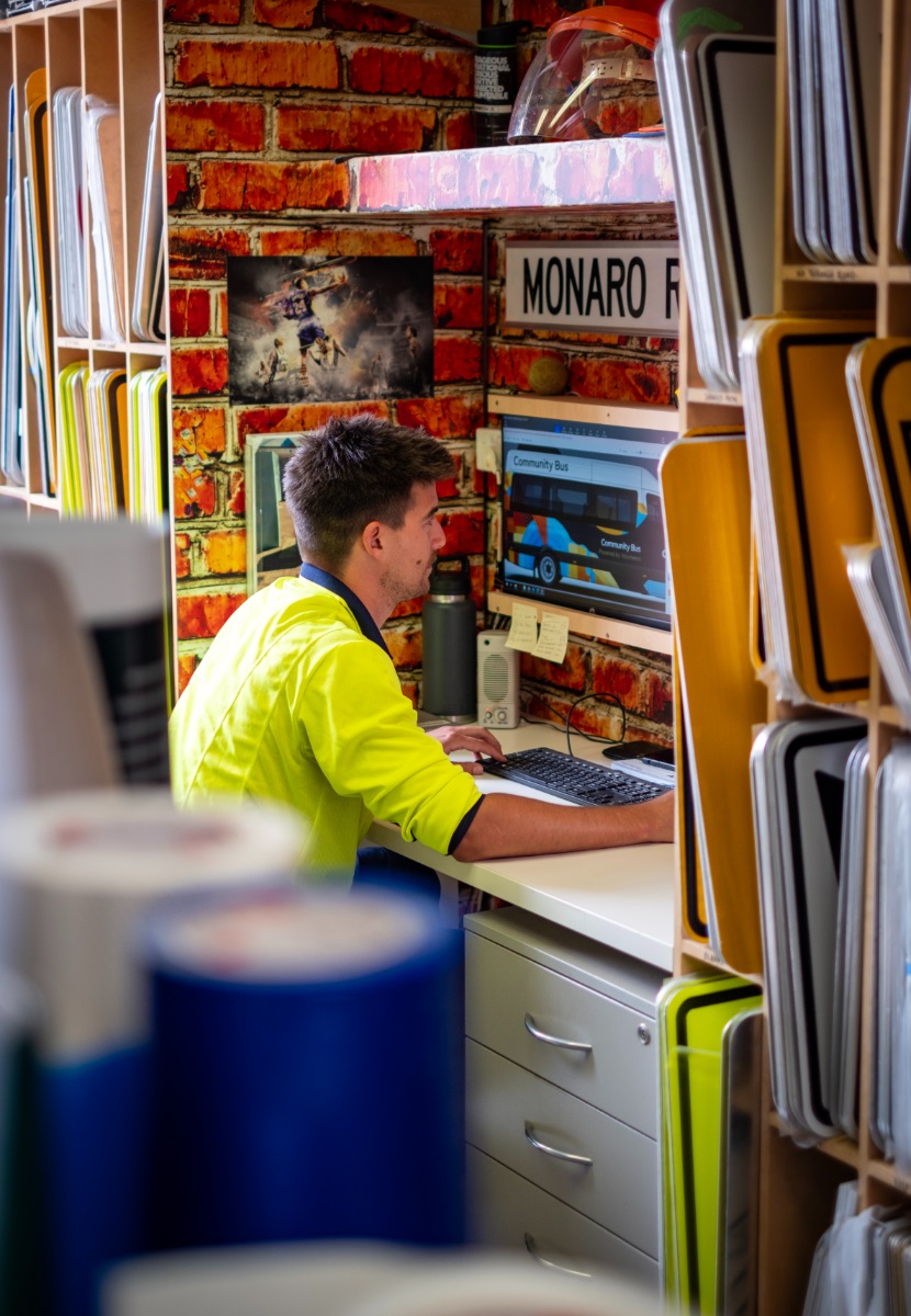 A worker in a high-vis shirt sits at a computer at the Sign Shop, surrounded by colourful signs.