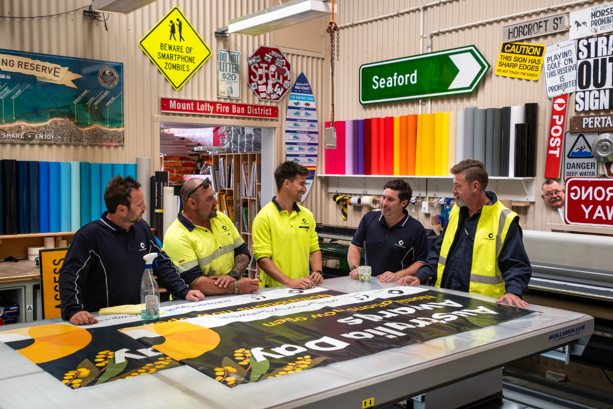 The five-person Sign Shop team chat over a table lined with signs, surrounded by colourful signage on the walls.