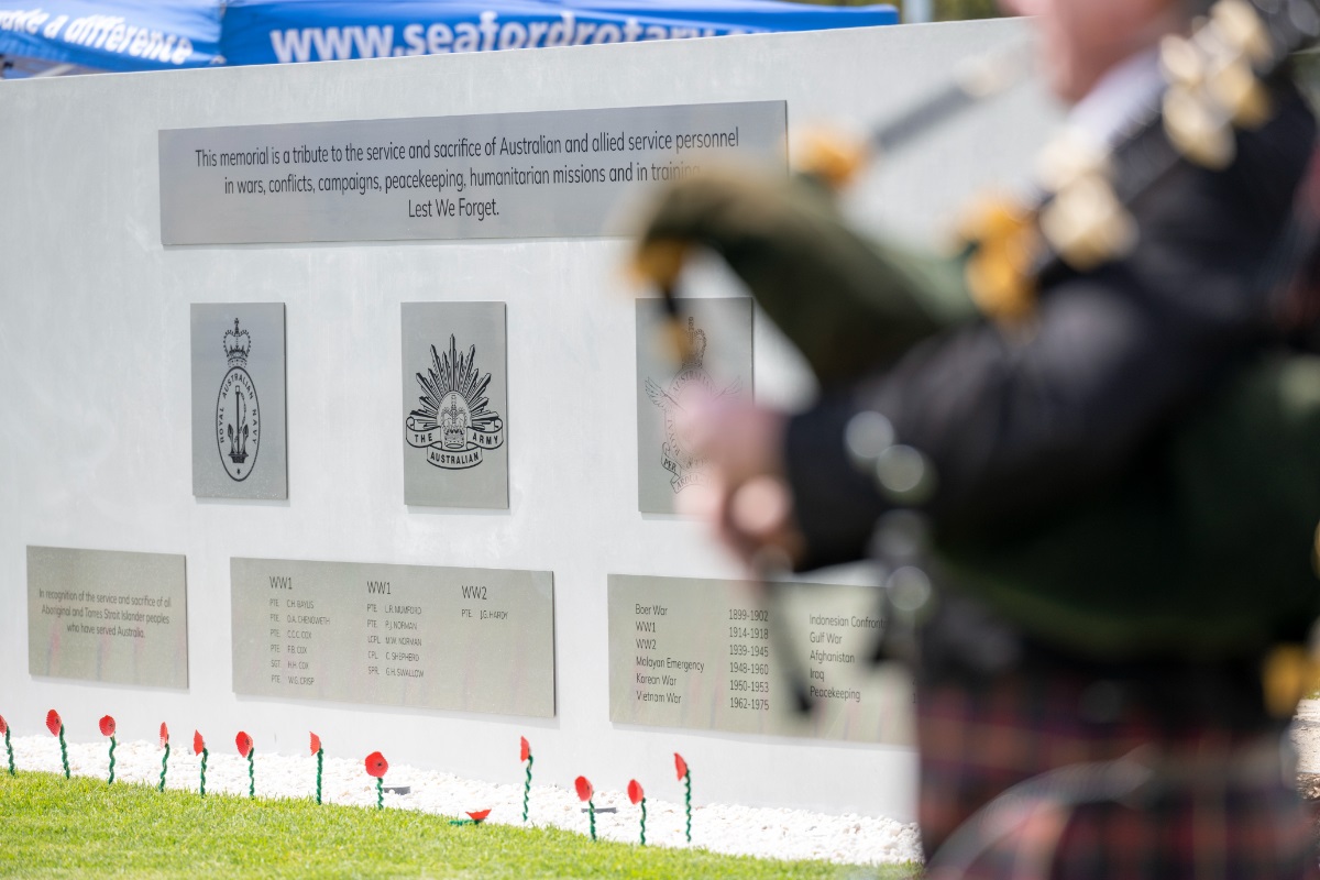 A bagpipe player, blurred in the foreground, stands in front of silver signage and poppies at a war memorial service.