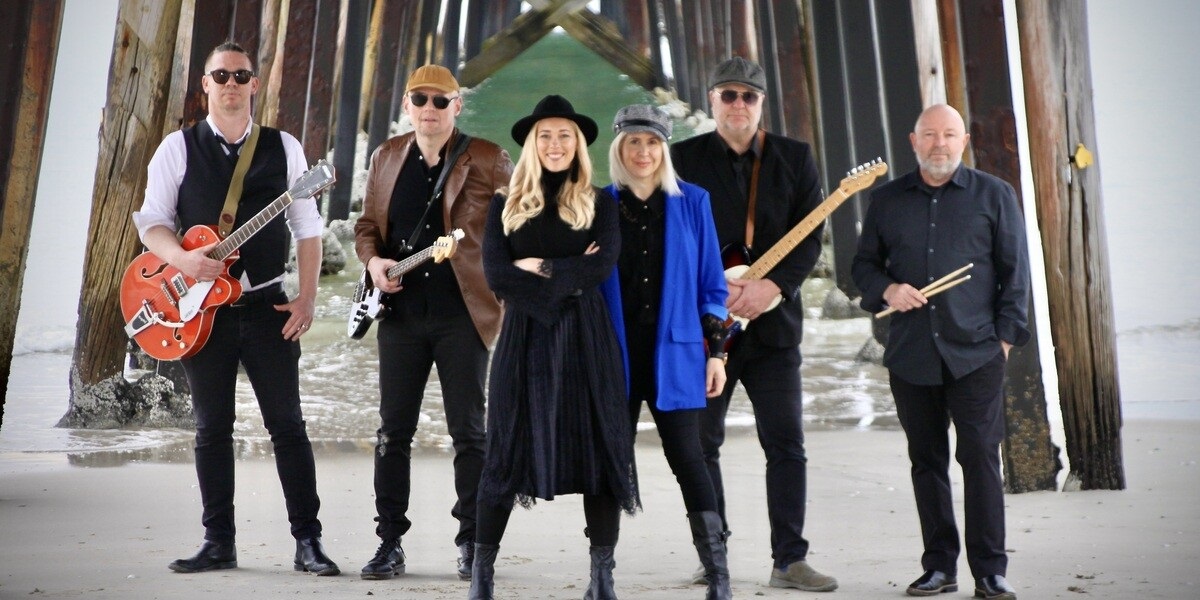 A band made up of two women in the centre, and three men holding instruments, stand underneath a jetty.
