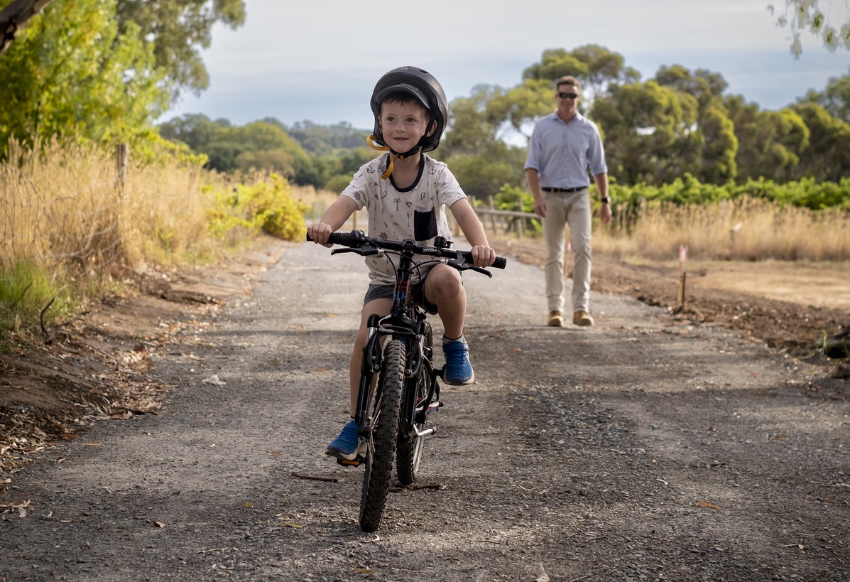 A boy smiles as he rides his bike along a rural trail with his father walking behind him.