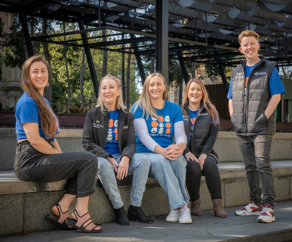 Five smiling people in blue Uniting Communities uniforms sit and stand at a set of stairs in an urban environment.