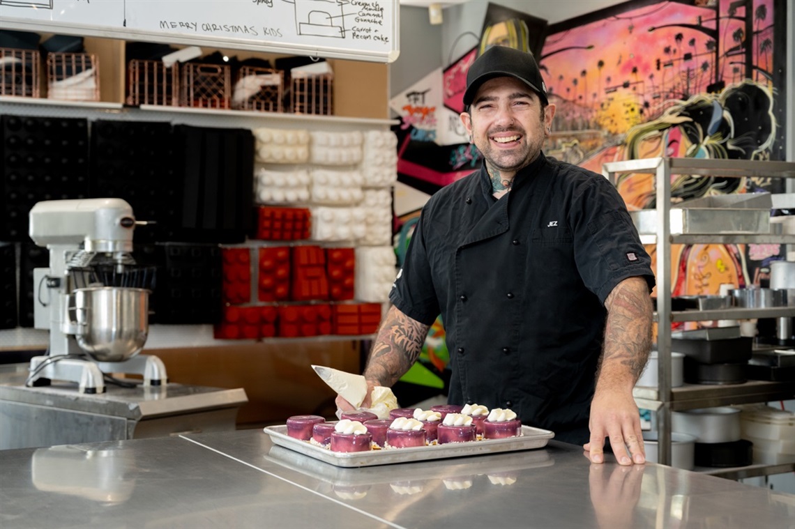 A smiling baker in his black uniform and black cap stands alongside a tray of deserts where he's half-way through topping them with cream.