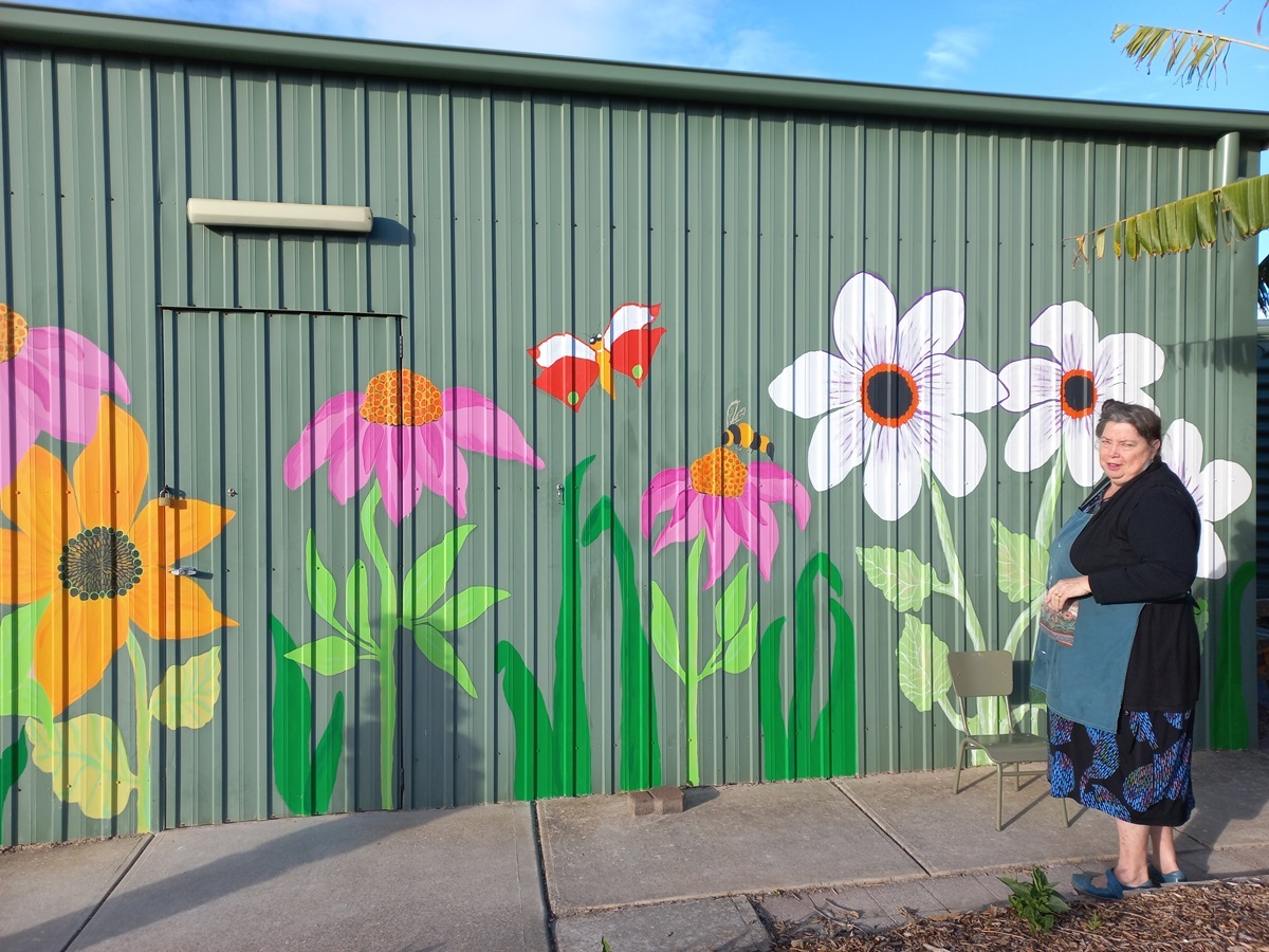 Volunteer Wendy Milano looks back at the camera alongside a beautifully painted shed exterior featuring bright flowers, a bee and a butterfly.