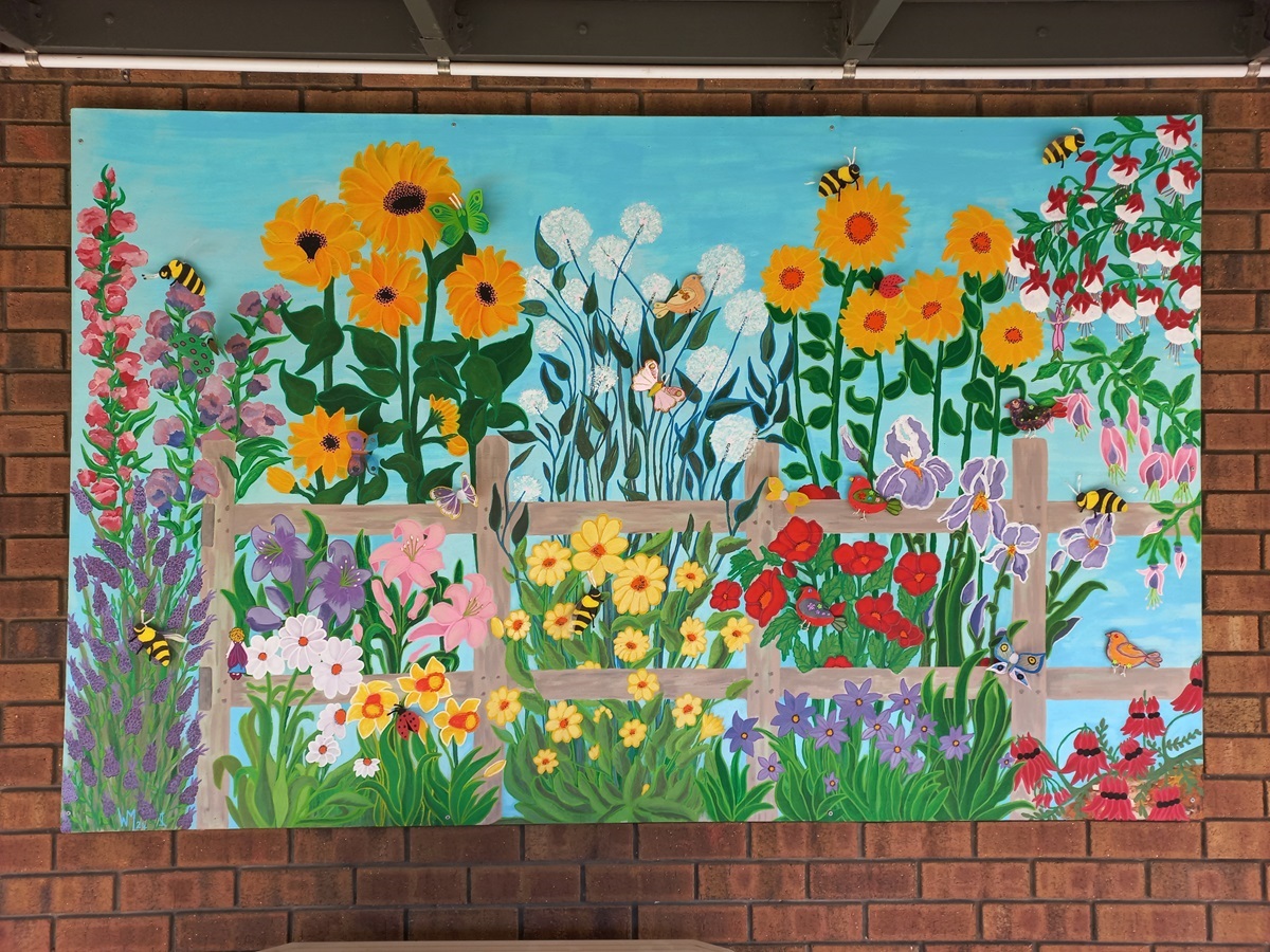 A large bright painting featuring a garden and pollinator scene hanging on a brick wall.