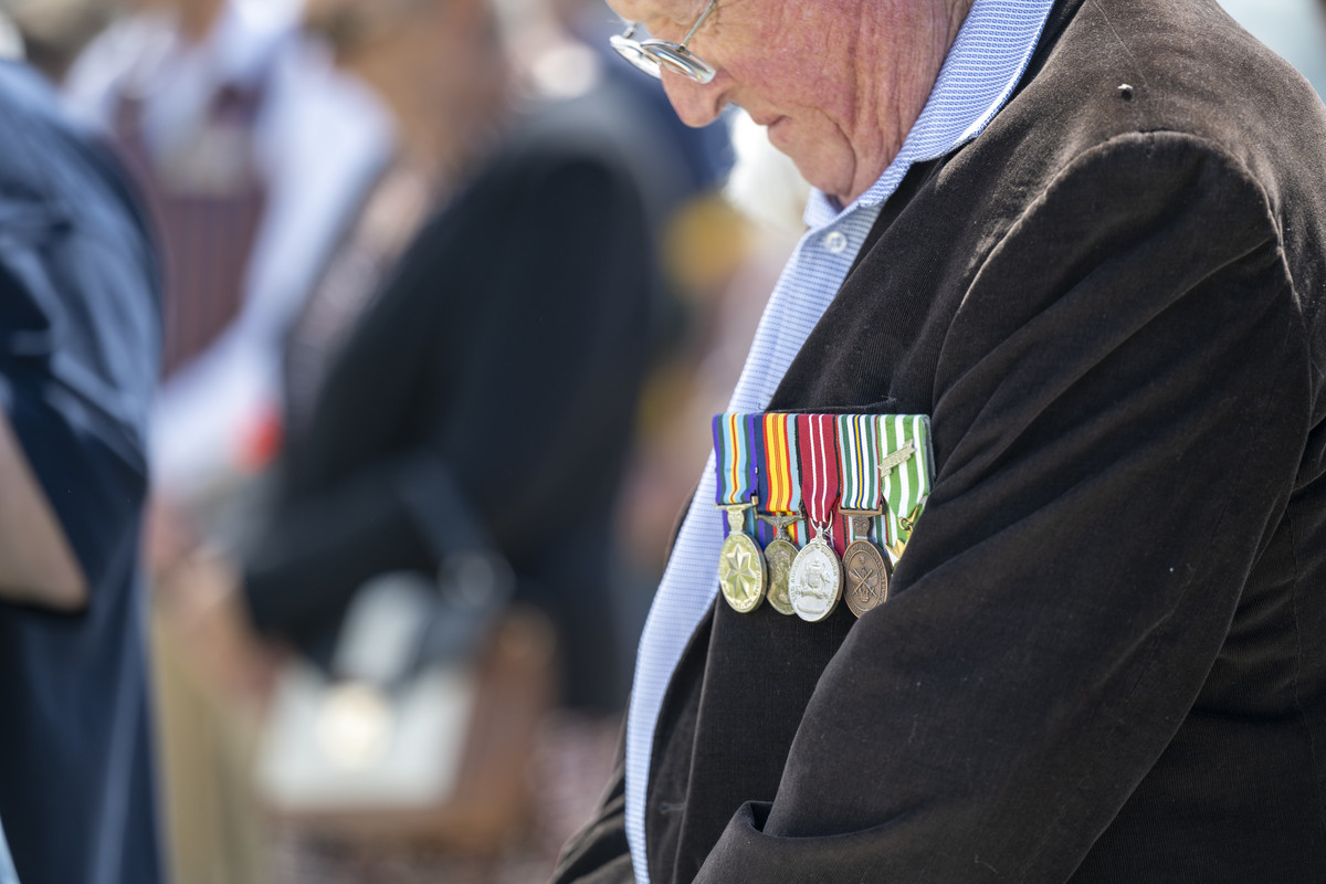 An elderly man wearing a row of war medals on the breast of his jacket bows his head during a commemorative war event.