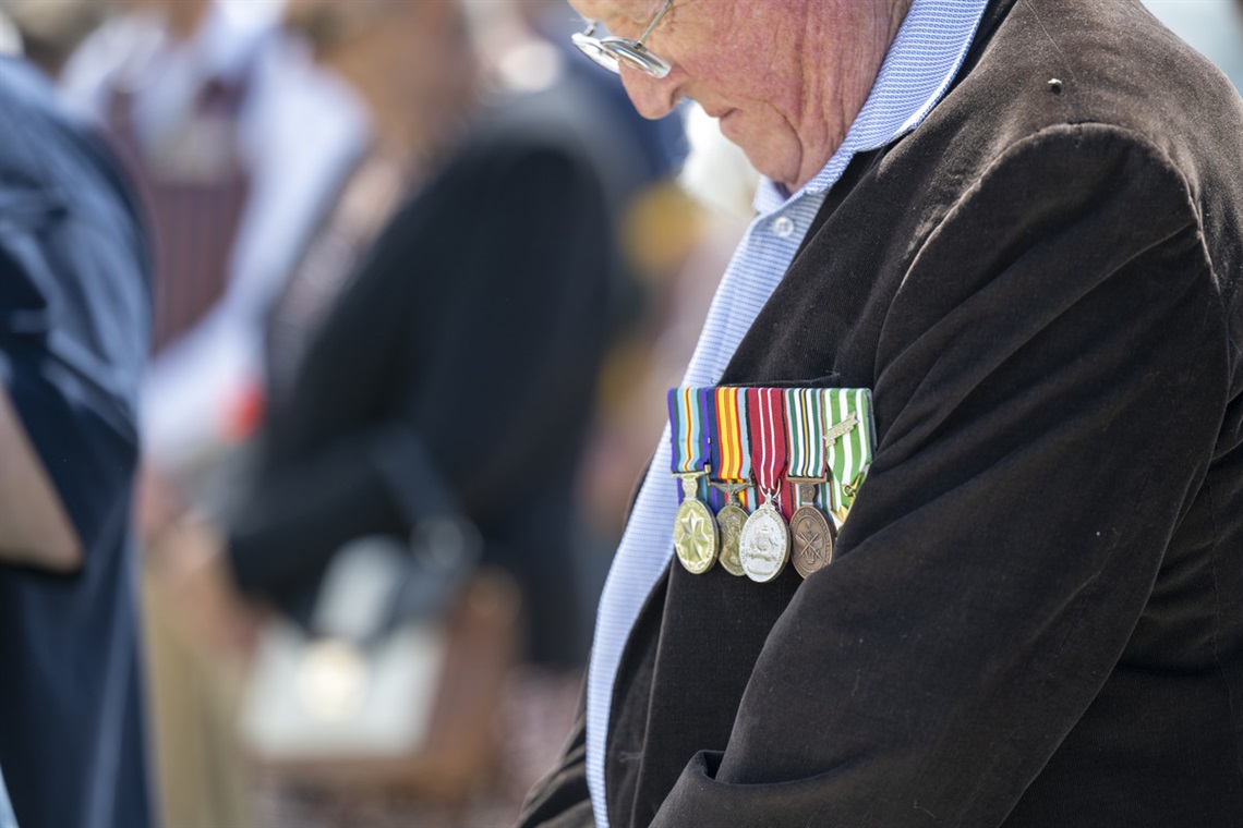 An elderly man wearing a row of war medals on the breast of his jacket bows his head during a commemorative war event.