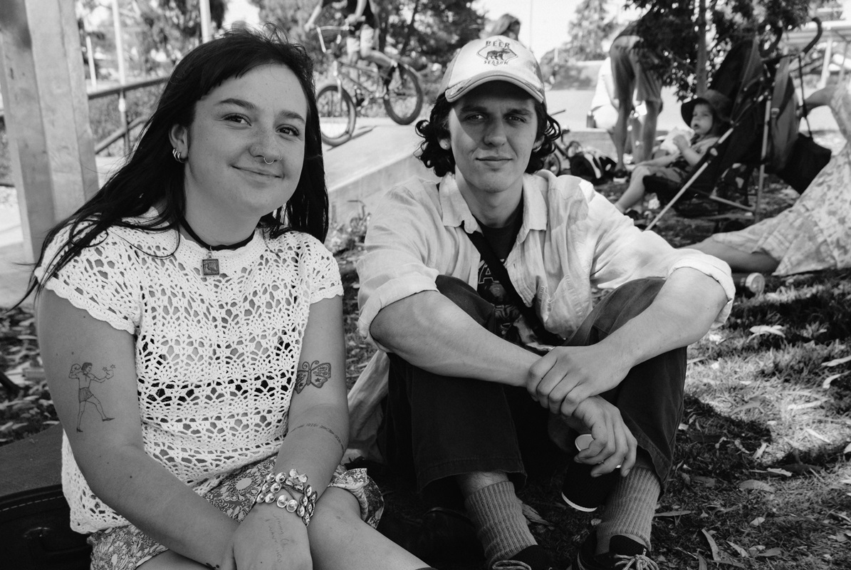 A black-and-white image of two smiling young people sitting together on grass with a skate park behind them.