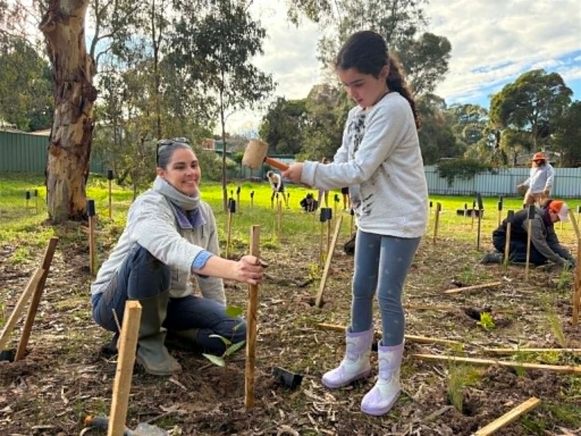 A mother holds a planting stake while her daughter hammers it in to the ground.