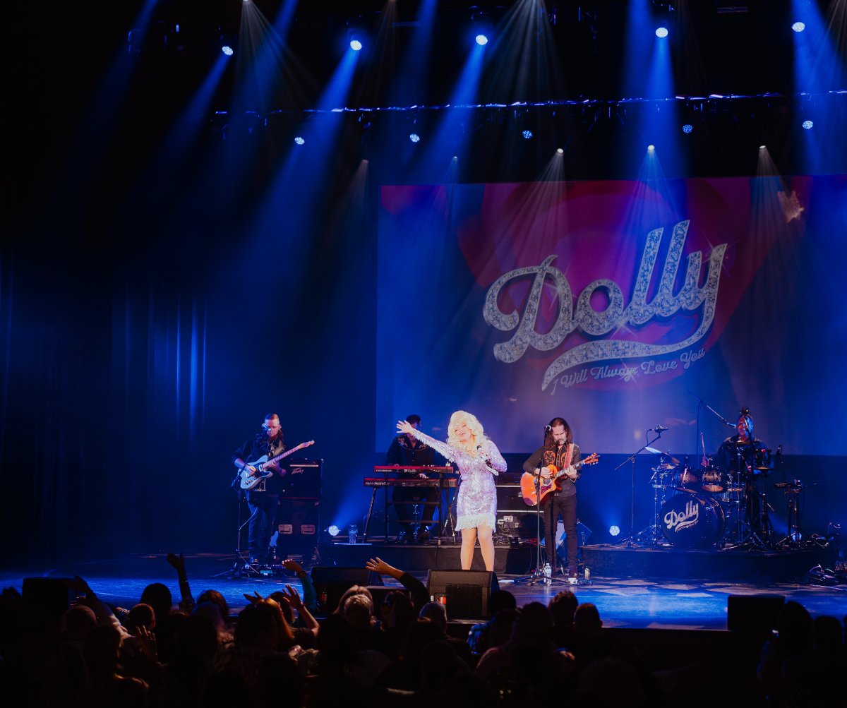 Dolly Parton tribute performer Brooke McMullen sings on stage with the words Dolly: I Will Always Love You on the wall behind stage.