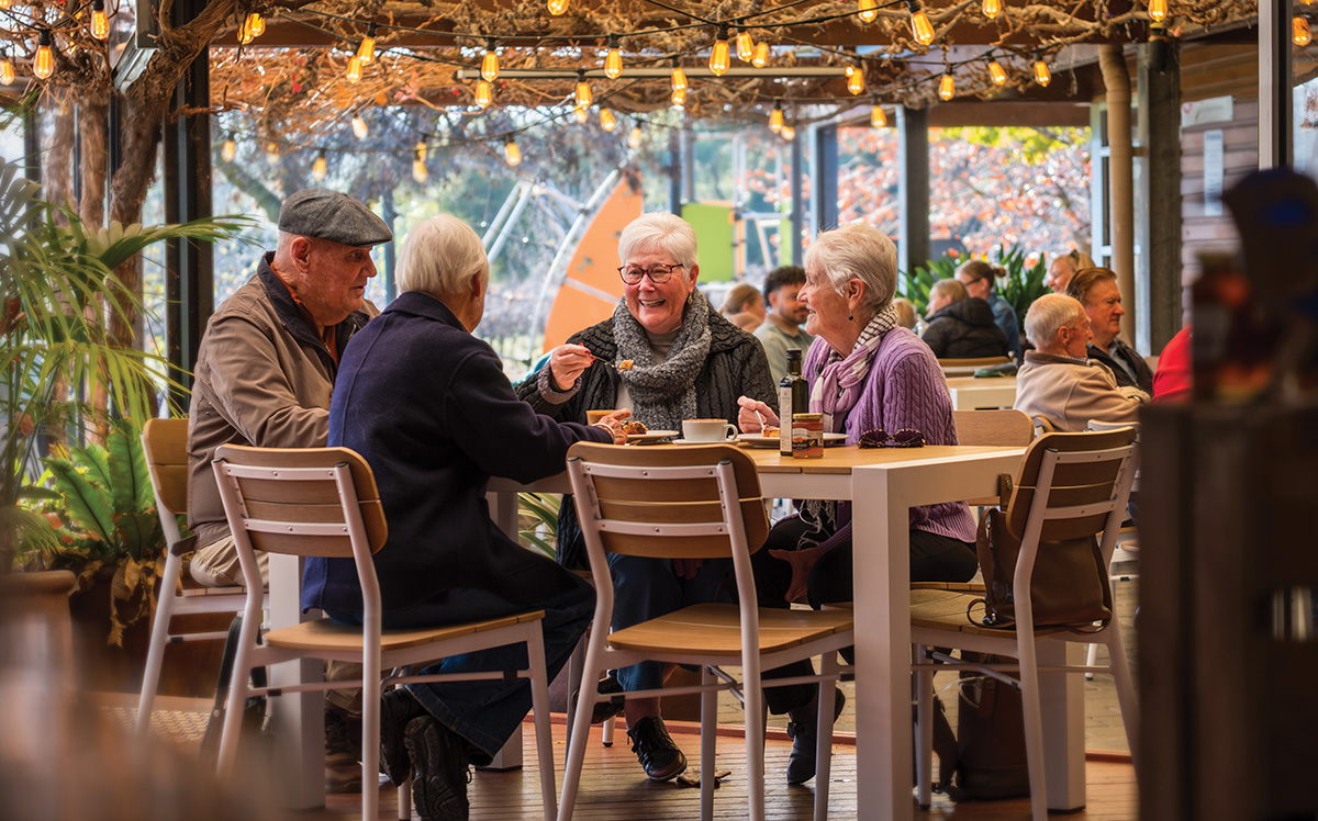 A group of older people sit around a table having morning tea. Fairy lights twinkle above them.