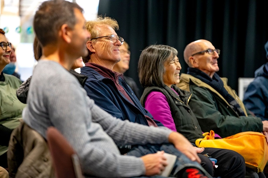 A crowd of smiling people sit watching a stage during an event.