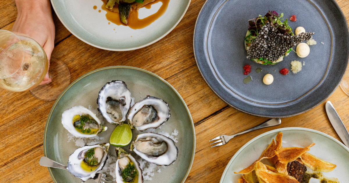 A timber table photographed from above featuring plates with oysters and other colourful gourmet food.