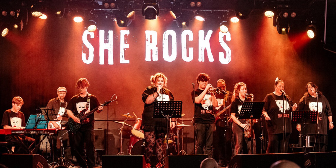 Musician Charlee Watt sings on stage alongside a band, with the words 'She rocks' behind them.