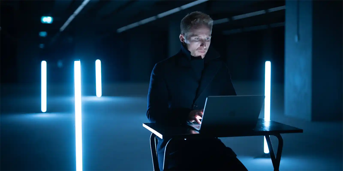 ABC host Hamish Mcdonald sits at a small black desk in a darkened room with strobe lights, typing at a laptop