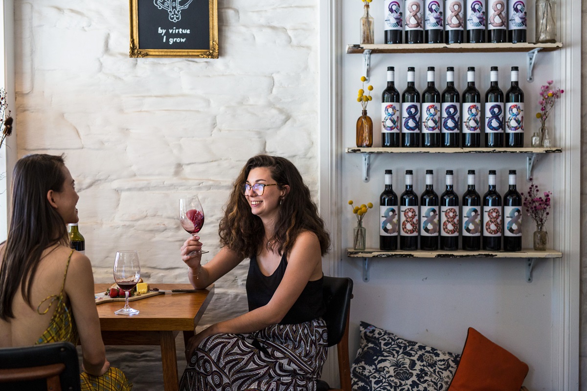 Two young people sit at a table in a winery with glasses of red wine. Shelves lined with red wine bottles sit alongside them.