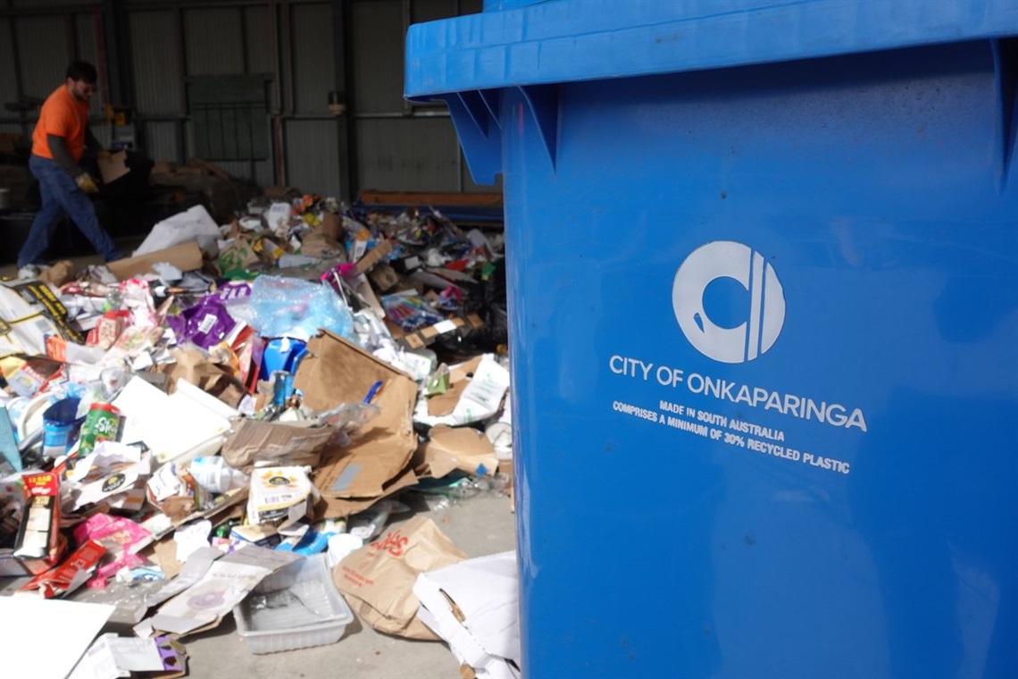 A worker in a high-vis orange vest inspects a pile of trash as part of a waste audit, with a blue City of Onkaparinga bin in the foreground.