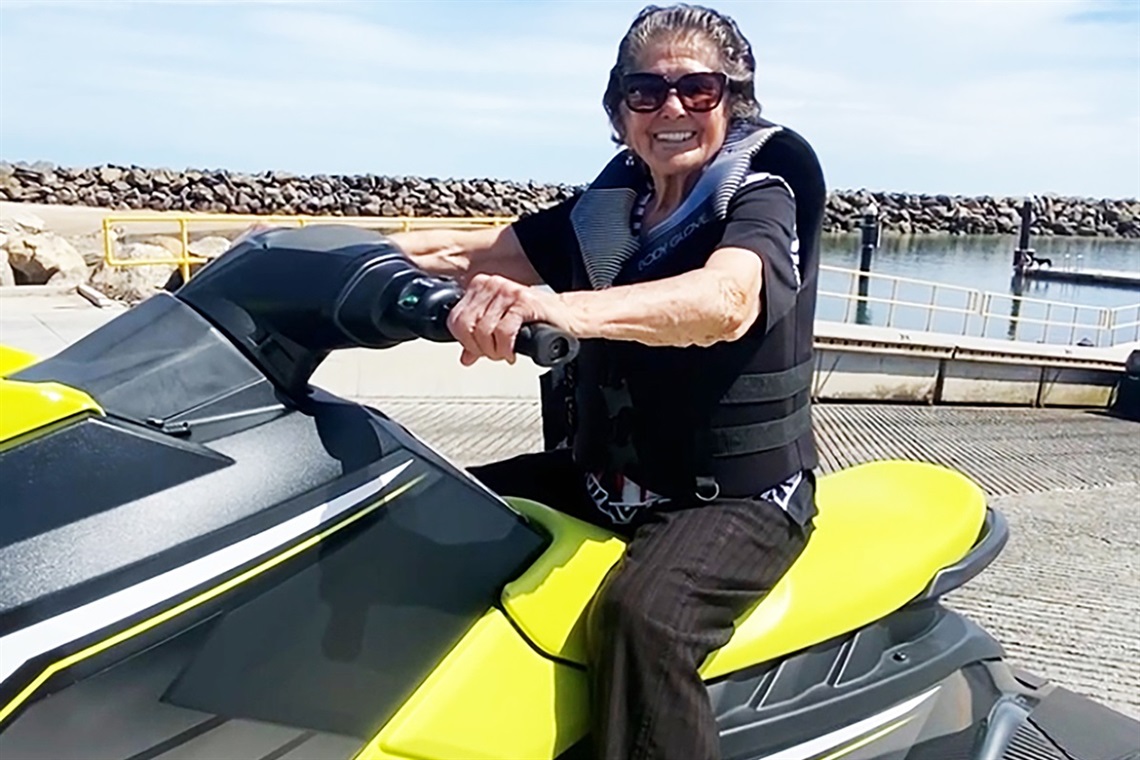 93-year-old Lola Gonzales smiles from atop a yellow-and-black jetski with a boat ramp in the background.