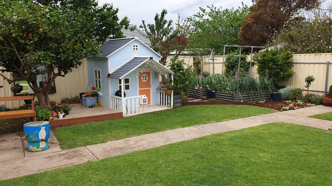 A well-manicured residential backyard garden featuring green lawns, a blue cubby house, veggie gardens and a large fruit tree.