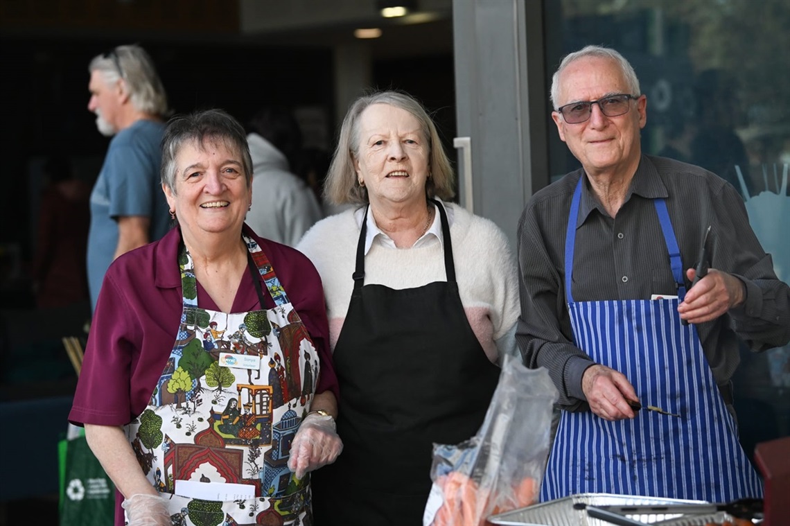 Three smiling older residents wearing aprons stand behind a barbecue at a community centre event.