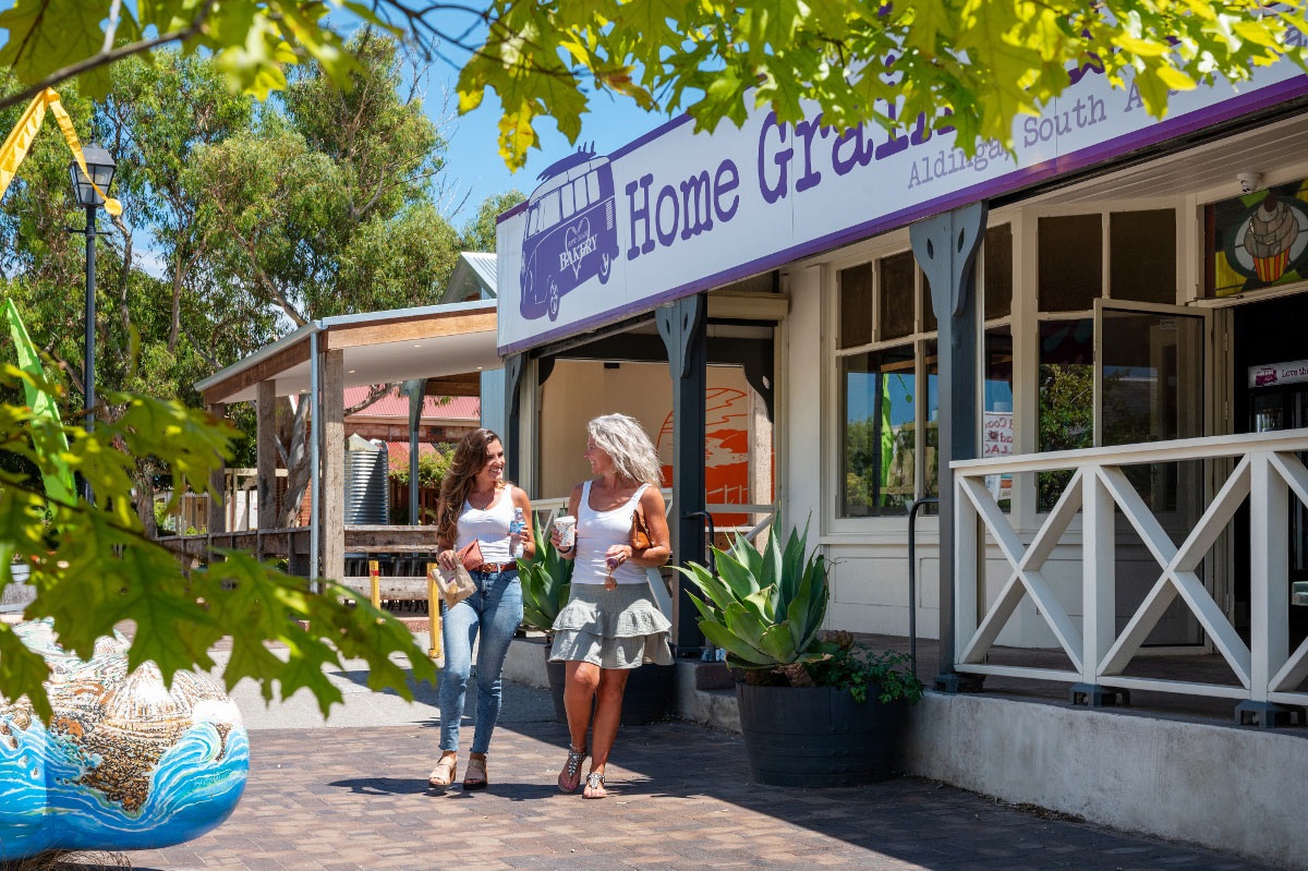 Two smiling women holding coffees walk past an Aldinga bakery.