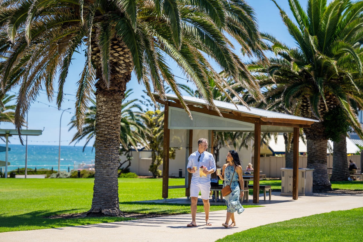 A smiling man and woman with coffees walk along a grass-lined path at Rotary Park, Christies Beach.