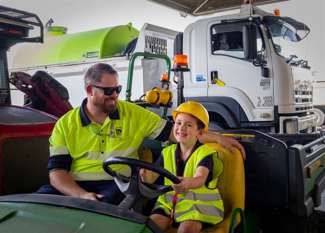 A smiling council worker in a high-vis shirt and sunglasses sits on a council buggy with a smiling boy in a high-vis vest and yellow helmet.