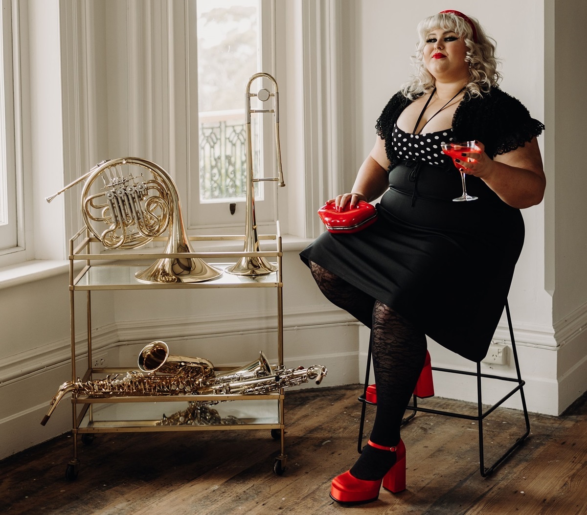 A photograph of musician Charlee Watt, who sits on a chair in a black dress and red lipstick in a sunlight-filled room, alongside shelves containing shiny horn instruments.
