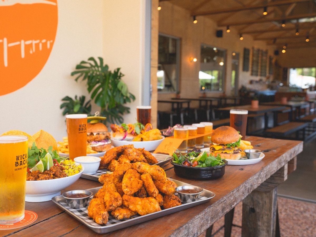Plates of stacked food including wings and burgers cover a wooden table at Kick Back Brewing.